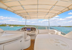 Cockpit view of a white luxury motorboat with beige seats, wooden deck, and a canopy, facing calm blue-green water and distant shoreline.