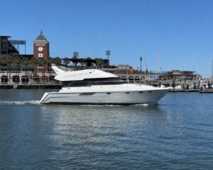 White motor yacht cruising in a calm harbor with a brick clock-tower building and palm-lined promenade in the background, under a clear blue sky.