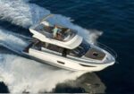 White motor yacht cruising fast on dark blue sea with two people on the upper deck on a sunny day.