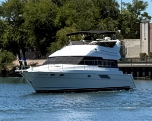 White motor yacht cruising on calm blue water near a shoreline with trees and a building in the background.