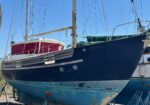 Side view of a dark blue sailboat hull in a dry dock with rigging and masts towering above, red cabin cover visible on the deck