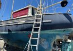 Blue sailboat out of the water on land with a metal ladder leaning against its hull and rigging above against a clear blue sky.