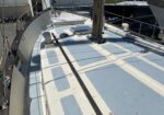 Deck of a sailboat with a blue-and-white non-slip pattern, central hatch, and standing rigging; other boats visible in the background at a marina.