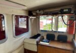 Interior view of a small boat cabin showing a wooden desk, beige upholstered walls, a mounted VHF radio and microphone, and a bright orange fire extinguisher on the right; windows reveal a marina outside.