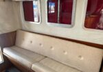 Interior of a vintage vehicle with a long beige upholstered bench seat and wood trim along the wall, next to red-tinted windows.