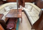Inside a boat cabin with cream cushioned seating around a wooden table and a metal pole in the center; gear and bags on the floor.