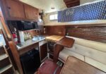 Compact boat galley with wooden cabinets, a stainless sink, microwave, and a fold-out table beside a cream upholstered bench.