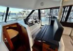 Cockpit of a luxury boat with helm, steering wheel, and navigation screens; seating to the right and cabin door open to a small cabin. Marina and yachts visible through windows.