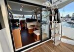 Open sliding glass door reveals a boat’s interior with a dining table, bench seating, and a ladder to the upper deck at a marina.
