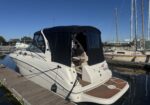 White motor yacht docked at a marina with a black canvas cover and wooden dock under a clear blue sky","Covered white boat tied to a wooden dock at a marina, masts in the background","Close-up of a white cabin cruiser with black canopy moored along a pier on calm water","Marina scene with a white motorboat in the foreground and sailboats with tall masts in the background","Boat at dock with protective black cover, calm water, blue sky