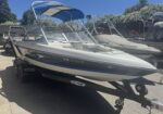 White and blue speedboat on a trailer in a sunny boatyard, other boats and flowering hedges in the background.
