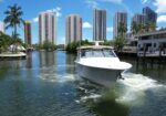White motorboat cruising through a canal with modern high-rise buildings and palm trees in the background by a blue sky.