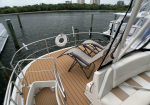A yacht deck with two loungers on a wooden surface, railings, and a life preserver, overlooking calm water and distant buildings.
