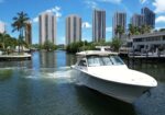 White yacht docked in a canal with palm trees and tall modern buildings in the background.