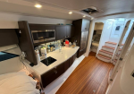 Interior of a modern yacht galley with dark wood cabinets, microwave, stovetop, and bottle bottles on a white countertop; wooden deck and curved stairs ahead.