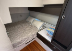 Compact cabin bedroom with a double bed, gray patterned duvet, and two pillows under built-in overhead storage in a boat or RV interior.
