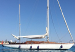 White sailing yacht with brown deck anchored on calm blue sea under a clear sky, sails furled and masts upright.