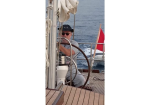 Older man wearing a hat and sunglasses steering a sailboat at the helm with rigging and a red flag in the background by the sea.