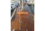 Teak deck of a sailing yacht at a marina, with ropes, rigging, and a hatch toward the bow.