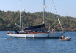 White sailing yacht anchored on blue water near a forested shoreline; several people on deck and a person in a small dinghy nearby.