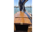 Teak-decked sailboat cockpit with an open storage hatch at the stern and rigging overhead, moored in a marina with several boats in the background.