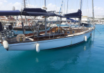 Sailing yacht docked in a marina with a white hull, wooden deck, blue awnings, and fenders along the side. A classic sailboat in calm turquoise water, moored at a harbor with rigging and lifebuoy visible on deck.