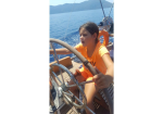 Girl in an orange shirt steering a sailboat, gripping the wheel with a bright ocean and distant mountains in view.