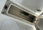 Open deck hatch on a white boat with dirt around the rim; inside are a green bag and a long cleaning tool/pole.