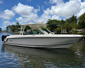 White motorboat docked on a calm canal with palm-lined shoreline and sunny tropical houses in the background, blue sky above.