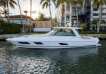 White luxury motor yacht docked on calm water with palm trees and apartment buildings in the background, sun glow behind the trees