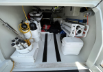 Boat storage compartment filled with gear: scuba tanks, yellow hoses, nets, folding equipment, cooler, and stray clothing/fabric items ready for use on deck.
