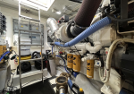 Engine room of a boat showing a large white diesel engine, blue hoses, and yellow Caterpillar fuel filters, with a metal ladder and maintenance gear nearby.