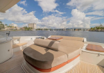 Rear deck of a luxury yacht with a curved beige sofa, teak decking, and small wooden tables, overlooking a waterway and city skyline under a blue sky.