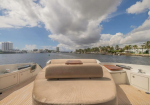 Luxurious boat deck with beige cushioned seating, teak floor, and built-in storage, facing a waterway with palm-lined shoreline and distant buildings under a cloudy sky