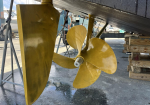 Large brass boat propeller under a hull, supported by stands in a marina/work yard.