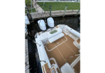 Top-down view of a small white boat docked at a wooden pier, with teak deck, seating area, and two raised outboard motors at the stern.