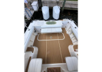 Top-down view of a white boat cockpit with tan teak deck, wraparound seating, and a center storage hatch at the rear by the docked area, with two white fenders nearby.