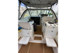 Interior of a small motorboat cabin with white cushioned seating, teak deck, helm at the front, and a compact galley with a sink on the right.