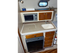 Compact boat galley with microwave, small sink, and a stovetop/oven beneath a beige countertop; wooden cabinets and drawers, porthole window above, and a fire extinguisher nearby.