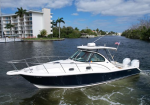 White and navy motor yacht cruising through a canal, with palm-lined waterfront buildings in the background and a small American flag at the stern.