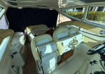 Interior of a beige yacht cabin with a captain's chair at the helm on the right, two rows of cushioned seats, wood trim, and large side windows.