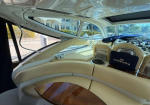 Cream leather lounge area inside a luxury boat, with curved seating around a wooden table and the helm up front; windows reveal a sunny marina outside.