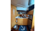 Compact boat galley with wood cabinetry, a two-burner stove and sink, upper glass-front cabinets, a small fridge, a blue anchor-themed rug on the deck floor, and a laundry basket nearby.