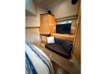 Compact boat cabin: bed on left, wooden cabinet and desk with a TV, and a black cushioned seating area with pillows.