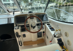 Boat cockpit with a wooden steering wheel, multiple navigation screens, and various controls, parked along a canal view through the windows.