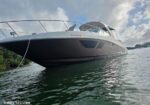 Large black-hulled motor yacht docked in calm water, viewed from near the bow with a railing and cabin area showing, shoreline greenery on the left and a partly cloudy blue sky above.
