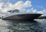 Grey motor yacht floating on calm water, bow angled toward the camera with railing and cabin visible under a blue cloudy sky