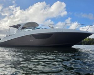 Grey motor yacht floating on calm water, bow angled toward the camera with railing and cabin visible under a blue cloudy sky