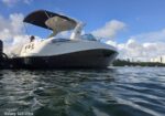 White motor yacht with a canopy on calm water, two people in the cockpit; city skyline on the horizon.