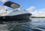 White luxury motor yacht on blue water with passengers on deck under a dark canopy and a city skyline in the distance